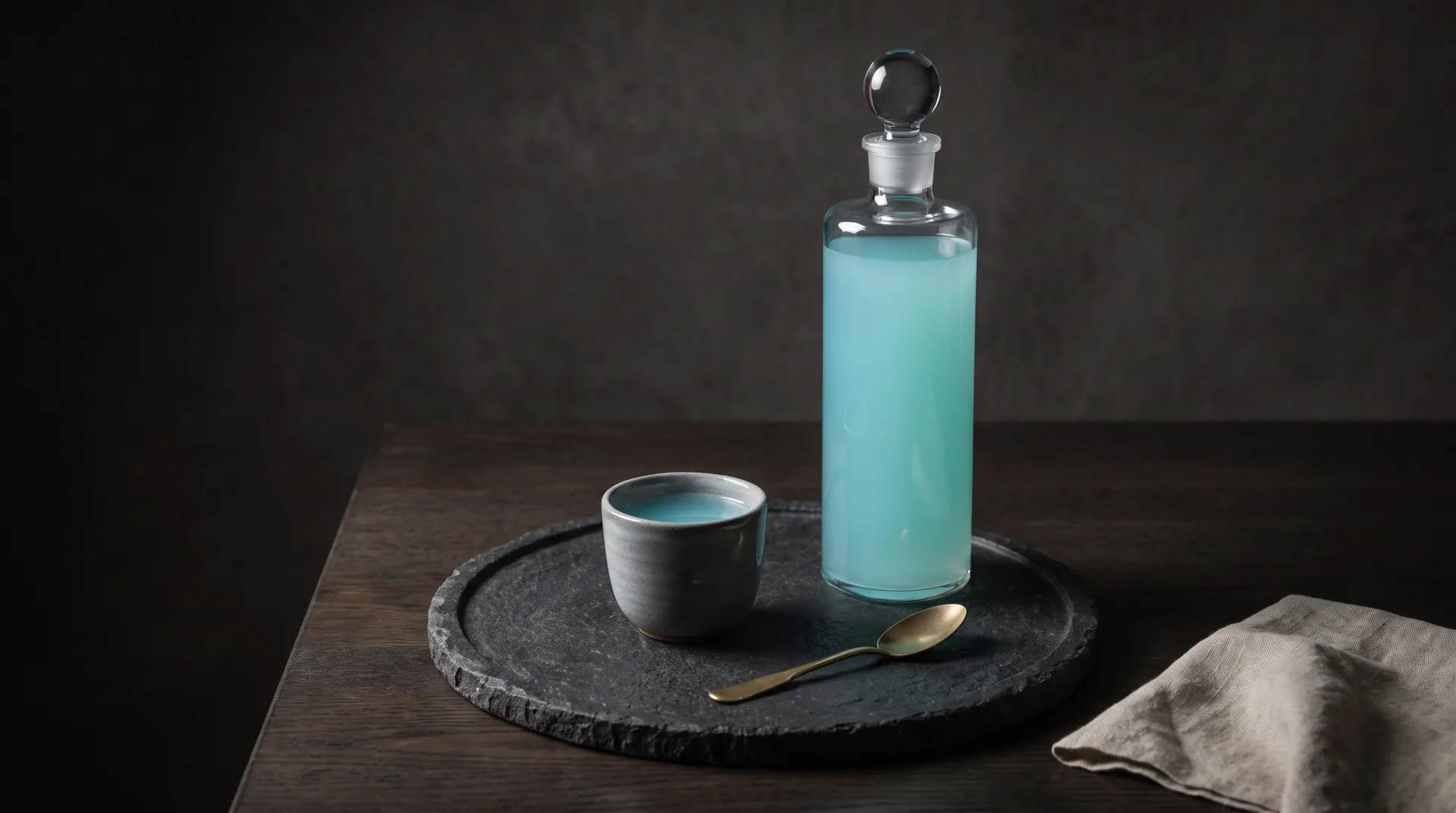 The bottle beside a stoneware cup and a brass spoon on a dark stone tray